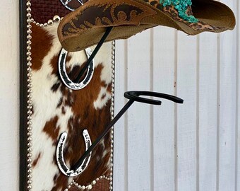 May include: Cowboy hat rack with a brown and white cowhide backing, adorned with silver horseshoe accents and a Dallas Cowboys star. The rack holds a brown straw cowboy hat with a decorative leather brim and turquoise accents.