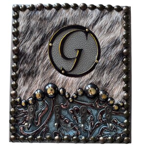 May include: A decorative key holder with a cowhide-like background and a large letter "G" in a circle. The key holder has four hooks and is framed with bronze-colored studs. The bottom section has a blue and brown floral design.