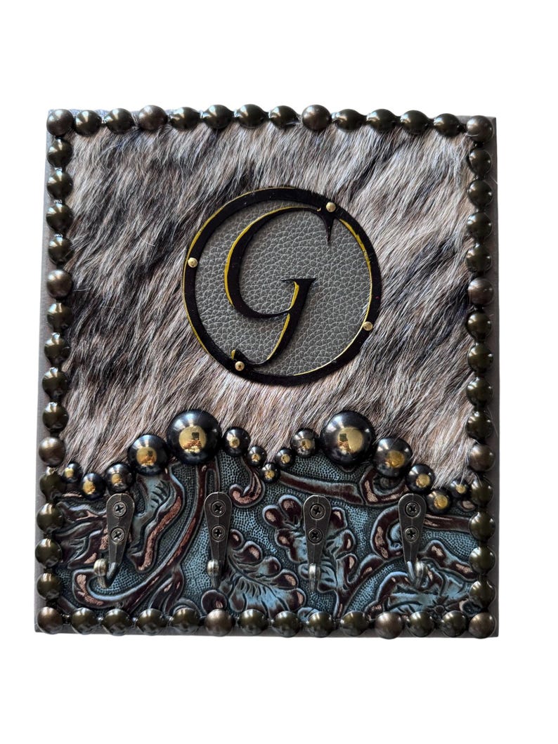 May include: A decorative key holder with a cowhide-like background and a large letter "G" in a circle. The key holder has four hooks and is framed with bronze-colored studs. The bottom section has a blue and brown floral design.