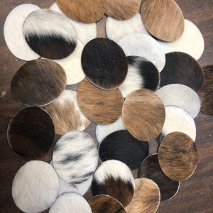 May include: A collection of oval-shaped coasters made from cowhide. The coasters display a variety of natural colors and patterns, including white, brown, black, and tan. The cowhide has a soft, textured appearance.
