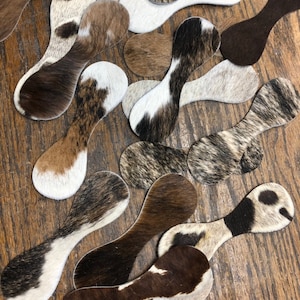 May include: A collection of cowhide spoon rests in various patterns and colors, including brown, white, and black. The spoon rests have a unique shape with a rounded end and a long, narrow handle. They are arranged on a wooden surface.