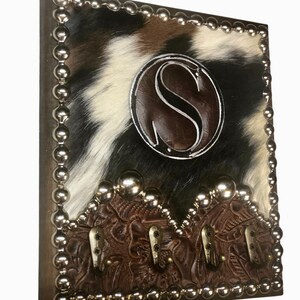 May include: A rustic key holder with a cowhide pattern in black, white, and brown. It features a brown leather accent with the letter "S" and decorative metal studs. Three metal hooks are attached to the bottom.