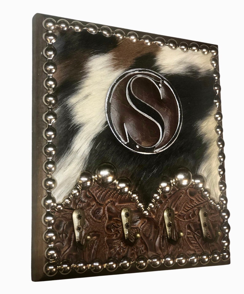 May include: A rustic key holder with a cowhide pattern in black, white, and brown. It features a brown leather accent with the letter "S" and decorative metal studs. Three metal hooks are attached to the bottom.