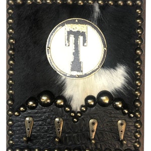 May include: A decorative wall-mounted coat rack with a cowhide background featuring black and white fur. The rack has a circular emblem with the letter "T" and four metal hooks. The frame is adorned with brass-colored studs.