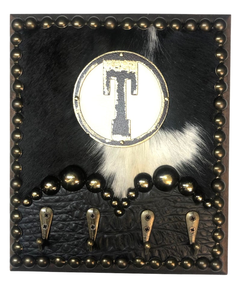 May include: A decorative wall-mounted coat rack with a cowhide background featuring black and white fur. The rack has a circular emblem with the letter "T" and four metal hooks. The frame is adorned with brass-colored studs.