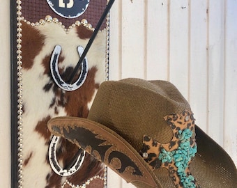 May include: A rustic cowboy hat rack with a brown and white cowhide design, a black horseshoe holder, and a brown straw cowboy hat with leopard print accents and turquoise stones. The rack has a woven leather top and decorative studs.