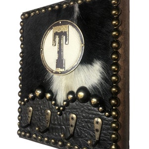 May include: A rustic wall-mounted key holder with a cowhide design. The holder features a black and white cowhide background, a circular emblem with the letter "T", and four metal hooks. The edges are adorned with brass-colored studs.