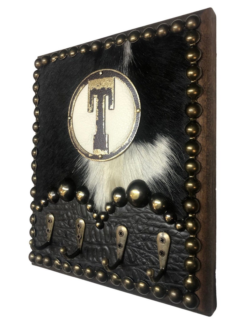 May include: A rustic wall-mounted key holder with a cowhide design. The holder features a black and white cowhide background, a circular emblem with the letter "T", and four metal hooks. The edges are adorned with brass-colored studs.