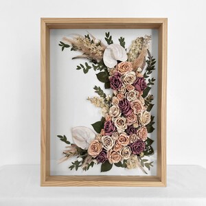 Pode incluir: Arranjo floral emoldurado com rosas em tons de rosa, p&ecirc;ssego e bord&ocirc;, callas brancas e gram&iacute;neas secas. O arranjo est&aacute; sobre um fundo branco dentro de uma moldura de madeira natural.