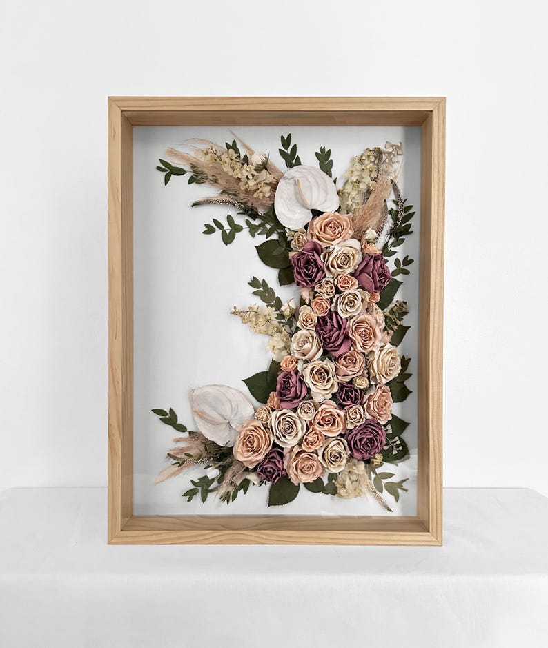 Pode incluir: Arranjo floral emoldurado com rosas em tons de rosa, p&ecirc;ssego e bord&ocirc;, callas brancas e gram&iacute;neas secas. O arranjo est&aacute; sobre um fundo branco dentro de uma moldura de madeira natural.