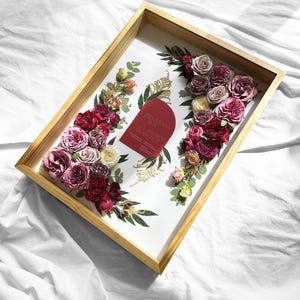 Pode incluir: Uma bandeja de madeira com interior branco, adornada com rosas bord&ocirc;, rosa e creme e vegeta&ccedil;&atilde;o. Um cart&atilde;o vermelho com texto dourado &eacute; colocado no centro. A bandeja est&aacute; sobre um fundo branco.