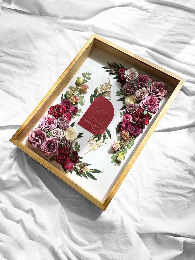 Pode incluir: Uma bandeja de madeira com interior branco, adornada com rosas bord&ocirc;, rosa e creme e vegeta&ccedil;&atilde;o. Um cart&atilde;o vermelho com texto dourado &eacute; colocado no centro. A bandeja est&aacute; sobre um fundo branco.