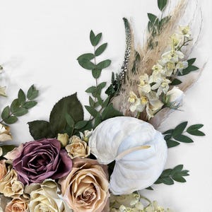 Pode incluir: Close-up de um arranjo floral com um ant&uacute;rio branco, rosas em tons de malva, p&ecirc;ssego e creme, e v&aacute;rias ervas secas e vegeta&ccedil;&atilde;o. O arranjo est&aacute; sobre um fundo branco, criando uma est&eacute;tica suave e natural.