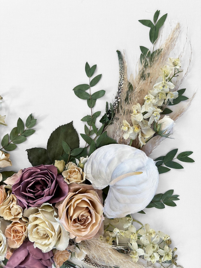 Pode incluir: Close-up de um arranjo floral com um ant&uacute;rio branco, rosas em tons de malva, p&ecirc;ssego e creme, e v&aacute;rias ervas secas e vegeta&ccedil;&atilde;o. O arranjo est&aacute; sobre um fundo branco, criando uma est&eacute;tica suave e natural.