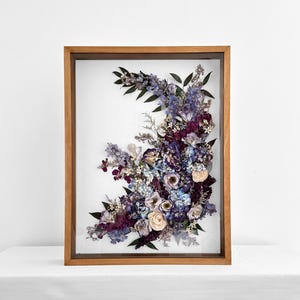 Pode incluir: Arranjo floral emoldurado com flores secas em tons de roxo, azul e creme. As flores s&atilde;o dispostas em forma semicircular dentro de uma moldura de madeira, criando uma pe&ccedil;a decorativa de arte de parede.