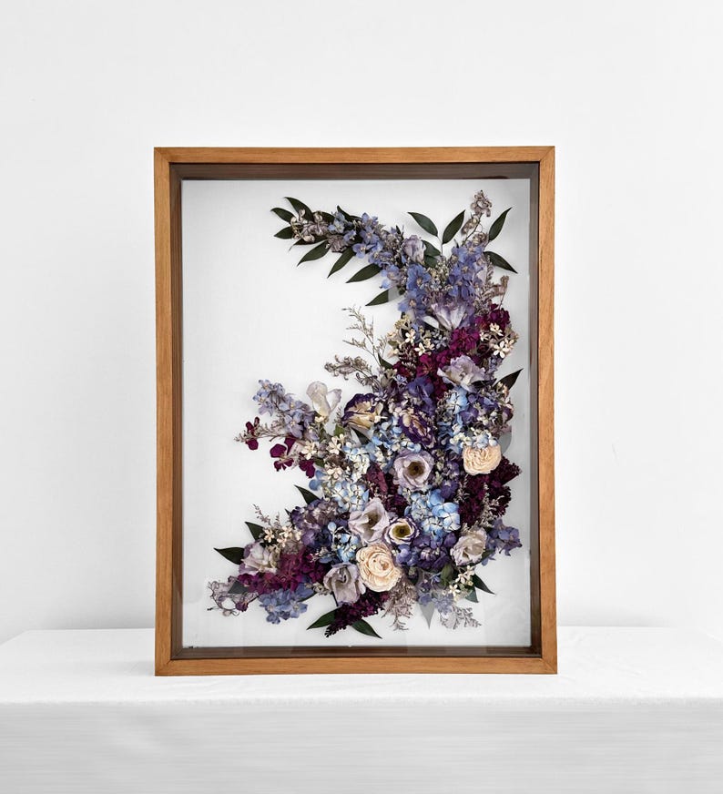 Pode incluir: Arranjo floral emoldurado com flores secas em tons de roxo, azul e creme. As flores s&atilde;o dispostas em forma semicircular dentro de uma moldura de madeira, criando uma pe&ccedil;a decorativa de arte de parede.