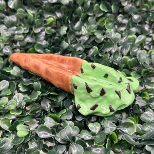 Può includere: Un cono gelato in ceramica con gelato verde menta e gocce di cioccolato. Il cono è marrone e ha una superficie strutturata. Il gelato è appoggiato su un letto di foglie verdi.