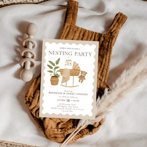 Peut inclure: Une invitation à une baby shower avec le texte "NESTING PARTY" présente une illustration à l'aquarelle d'un berceau, d'un ours en peluche et d'une plante en pot. L'invitation est sur un fond beige, avec un body marron et un hochet en bois en arrière-plan.