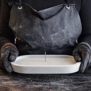 Könnte beinhalten: Eine hellgraue, ovale Betonschale mit einem einzelnen, aufrechten Metallstift in der Mitte. Die Schale wird von einer Person mit schwarzen Handschuhen und einer Schürze gehalten. Der Hintergrund ist dunkel.