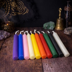 Peut inclure: Une collection de dix bougies coniques minces et colorées disposées sur une surface en bois. Les bougies sont dans un spectre de couleurs arc-en-ciel, comprenant le violet, le bleu, le rose, le jaune, l'orange, le rouge, le vert, le noir et le blanc. Chaque bougie a une mèche blanche.