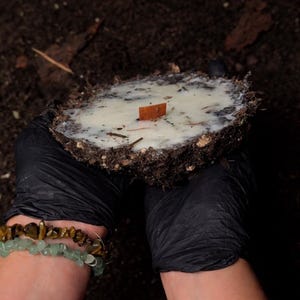 Puede incluir: Una vela hecha a mano con una estética rústica. La vela es circular, con una superficie de cera blanca cremosa y un pequeño acento de madera rectangular. El borde exterior está compuesto de tierra y materia orgánica. La vela es sostenida por una persona con guantes negros.