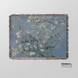 May include: A woven throw blanket featuring a reproduction of Vincent van Gogh's painting "Almond Blossom." The painting depicts a white almond tree in full bloom against a blue sky.