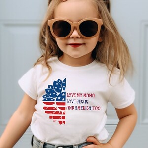 May include: A white t-shirt with a red, white, and blue sunflower design. The sunflower is made up of the American flag. The text on the shirt reads "Love my mama, love Jesus, and America too."