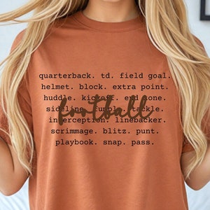 May include: A rust-colored t-shirt with a brown graphic print of the word "football" and a list of football terms. The text reads: "quarterback. td. field goal. helmet. block. extra point. huddle. kickoff. end one. football sideline fumble tackle. interception. linebacker. scrimmage. blitz. punt. playbook. snap. pass."