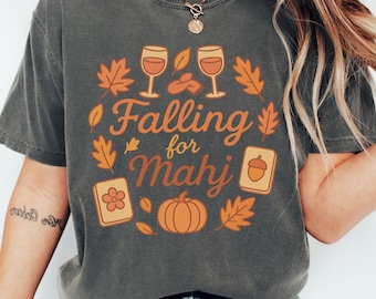May include: Dark gray t-shirt with a fall-themed graphic. The design includes two wine glasses, autumn leaves, a pumpkin, and the text "Falling for Mahj". The shirt is a comfortable, casual style.