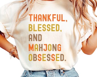 May include: Cream-colored t-shirt with the text "THANKFUL, BLESSED, AND MAHONG OBSESSED" in orange, yellow, and brown lettering. The shirt is made of a soft material and is being worn with blue jeans.
