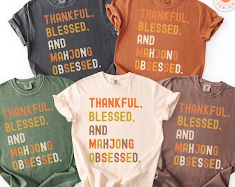 May include: Five different colored t-shirts with the text "THANKFUL, BLESSED, AND MAHJONG OBSESSED." The shirts are in shades of gray, orange, green, cream, and brown. The text is in a mix of orange, yellow, and brown.