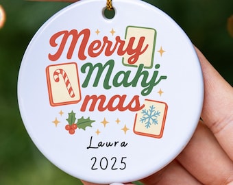 May include: A white ceramic Christmas ornament with the words "Merry Mahj mas" in red and green, along with a candy cane, snowflake, and holly illustrations. The name "Laura" and the year "2025" are printed below.