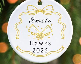 May include: White ceramic Christmas ornament with gold accents. The ornament features the name "Emily", the text "Hawks 2025", and a design of two bells with a bow. A gold string is attached for hanging.