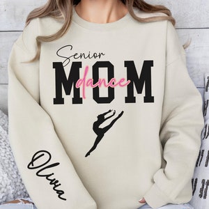 May include: A beige sweatshirt with the text "Senior Dance Mom" in black with a pink "dance" and a silhouette of a dancer. The left sleeve has the name "Olivia" written in black.