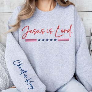 May include: Gray sweatshirt with the text "Jesus is Lord." in red lettering and a red, white, and blue American flag design. The left sleeve has the text "Christ is King." in blue lettering.