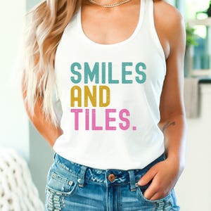May include: White tank top with the words "SMILES AND TILES" in teal, gold, and pink block letters. The top is paired with blue denim jeans. The overall style is casual and comfortable.