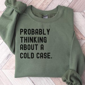 May include: A green sweatshirt with the text "PROBABLY THINKING ABOUT A COLD CASE." printed in black.