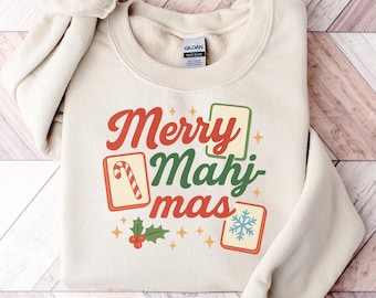 May include: Cream-colored sweatshirt with the text "Merry Mahjmas" in red and green. The design includes festive icons like a candy cane, snowflake, and holly. The sweatshirt is folded on a wooden surface.
