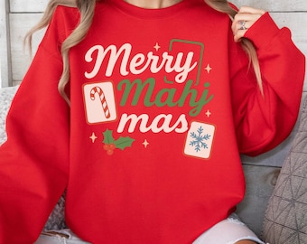 May include: A red sweatshirt with the words "Merry Mahj Mas" in white and green lettering. The design includes a candy cane, snowflake, and holly sprig. The sweatshirt is a festive holiday garment.