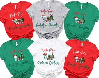 May include: Six t-shirts in different colors with a Christmas-themed design featuring a tooth wearing a Santa hat and holding a gift box and a Christmas tree. The text on the shirts reads "North Pole Pediatric Dentistry".