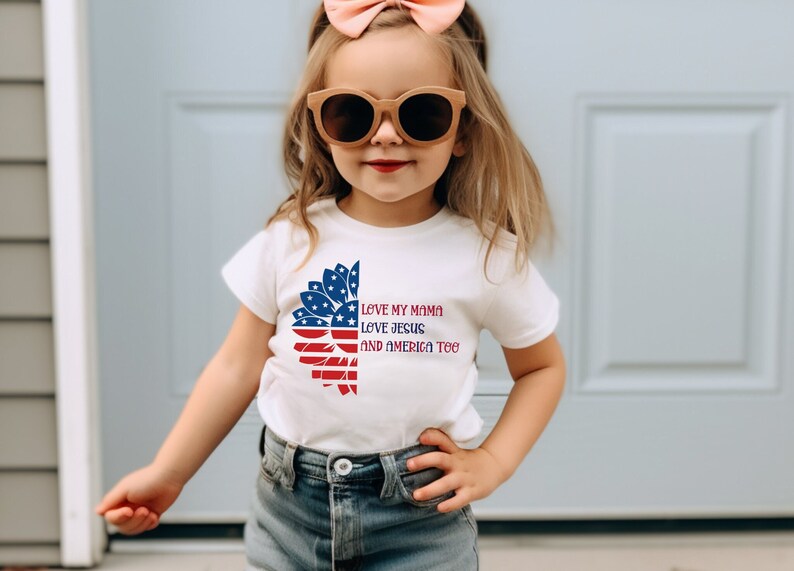 May include: A white t-shirt with a red, white, and blue sunflower design. The sunflower is made up of the American flag. The text on the shirt reads "Love my mama, love Jesus, and America too."