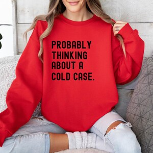 May include: Red sweatshirt with the text "PROBABLY THINKING ABOUT A COLD CASE." printed in black.
