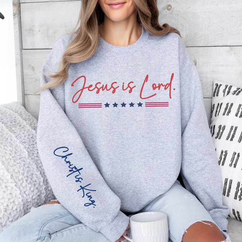 May include: Gray sweatshirt with the text "Jesus is Lord." in red lettering and a red, white, and blue American flag design. The left sleeve has the text "Christ is King." in blue lettering.