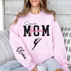 May include: Pink sweatshirt with the text "Senior Dance Mom" in black and pink lettering. The sweatshirt has a silhouette of a dancer on the front and the name "Olivia" on the sleeve.