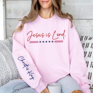 May include: Pink sweatshirt with the text "Jesus is Lord." in red and "Christ is King." in blue on the sleeve. The sweatshirt has a red, white, and blue stripe with five stars.