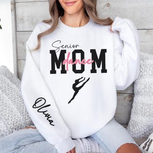 May include: White sweatshirt with the text "Senior dance MOM" in black with a pink "dance" and a silhouette of a dancer. The left sleeve has the name "Olivia" written in black.