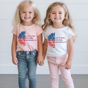 May include: Two young girls wearing matching t-shirts with a sunflower design and the text "Love my mama, love Jesus, and America too". The girl on the left is wearing a pink shirt and blue jeans, while the girl on the right is wearing a white shirt and pink pants.
