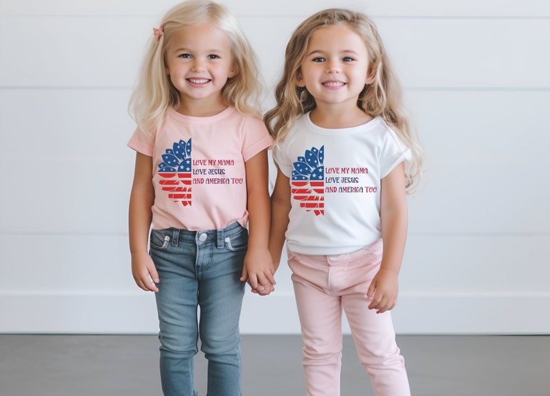 May include: Two young girls wearing matching t-shirts with a sunflower design and the text "Love my mama, love Jesus, and America too". The girl on the left is wearing a pink shirt and blue jeans, while the girl on the right is wearing a white shirt and pink pants.