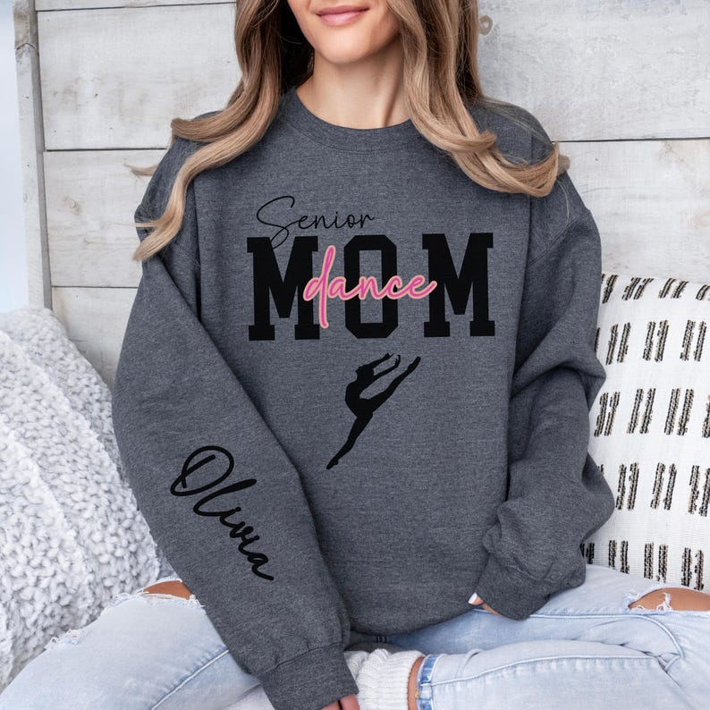 May include: Gray sweatshirt with the text "Senior dance MOM" in black with the word "dance" in pink. The sweatshirt has a silhouette of a dancer on the front and the name "Olivia" on the left sleeve.