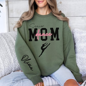 May include: A green sweatshirt with the text "Senior Dance Mom" in black and pink lettering. A silhouette of a dancer is below the text. The word "Olivia" is written in cursive on the left sleeve.